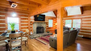 Et interiør i en bjælkehytte med en stenvægge og pejs, et træspisebord med stole, en sofa og et skilt med teksten 'Smoky Mountain Stays'.