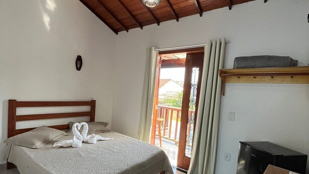 A bedroom with a bed, a desk, a refrigerator, and a balcony door. - Pousada Luz da Lua (Búzios)