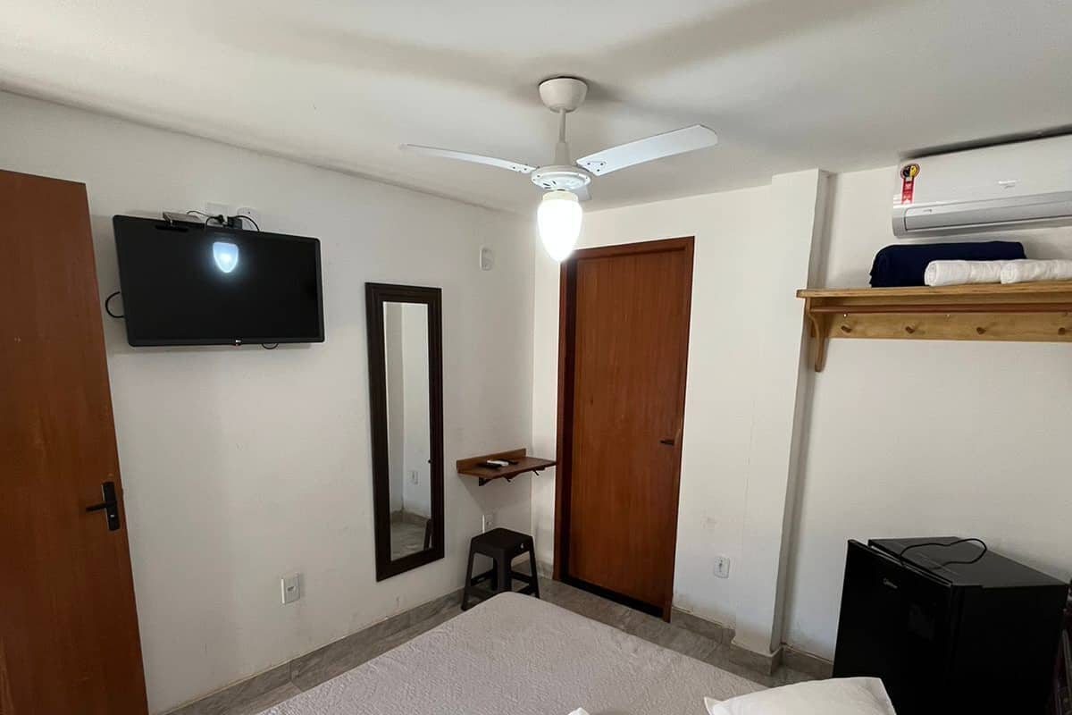 A bedroom with a bed, a TV mounted on the wall, a ceiling fan, and a wooden shelf with a heater.