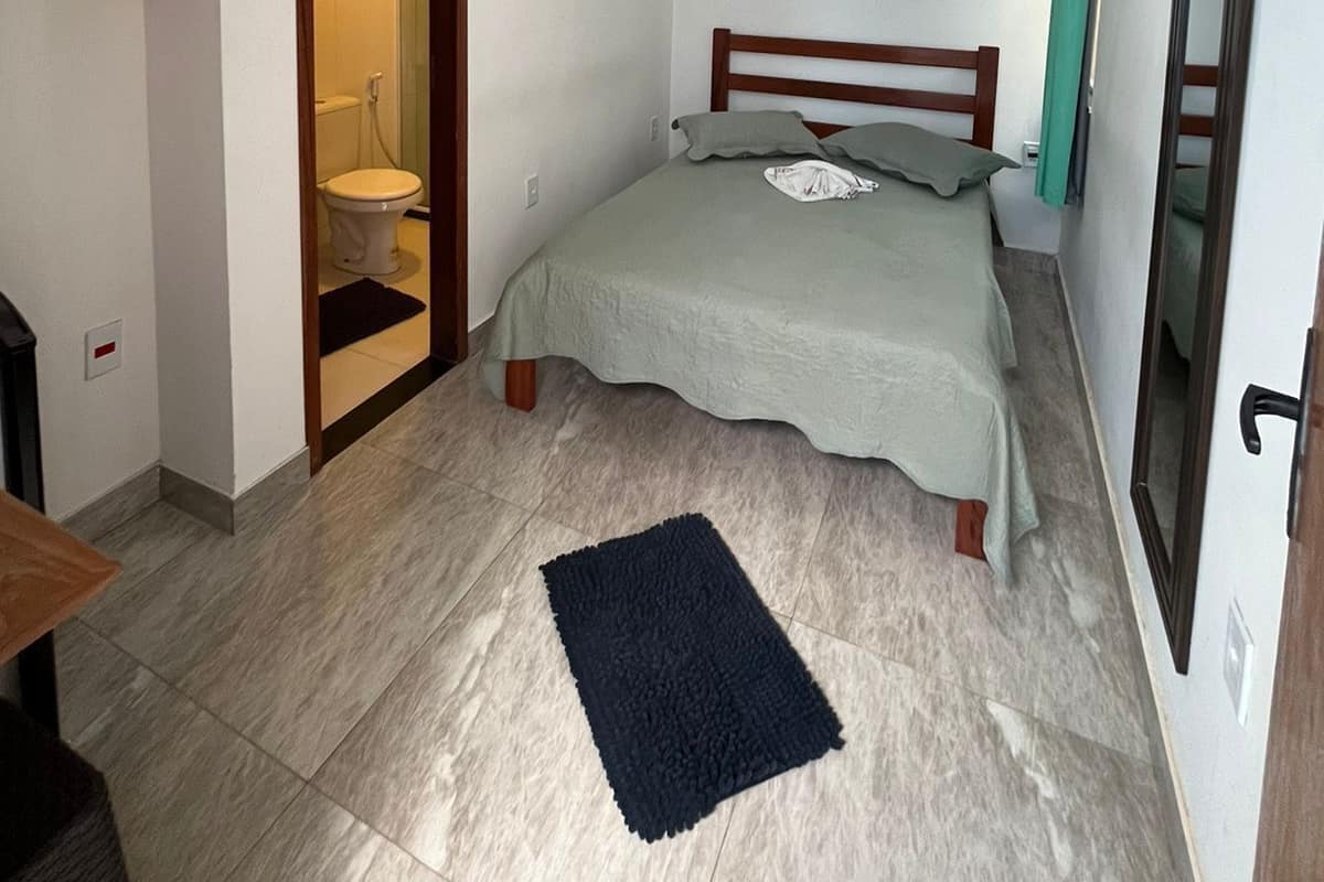 A small, well-lit bedroom with a single bed, a ceiling fan, and a visible bathroom door.