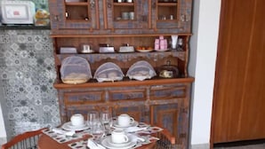 A dining area with a round table set for four, surrounded by four chairs with metal frames and wooden seats. Above the table, there is a wooden cabinet with glass doors containing various kitchen items. - Pousada Luz da Lua (Búzios)