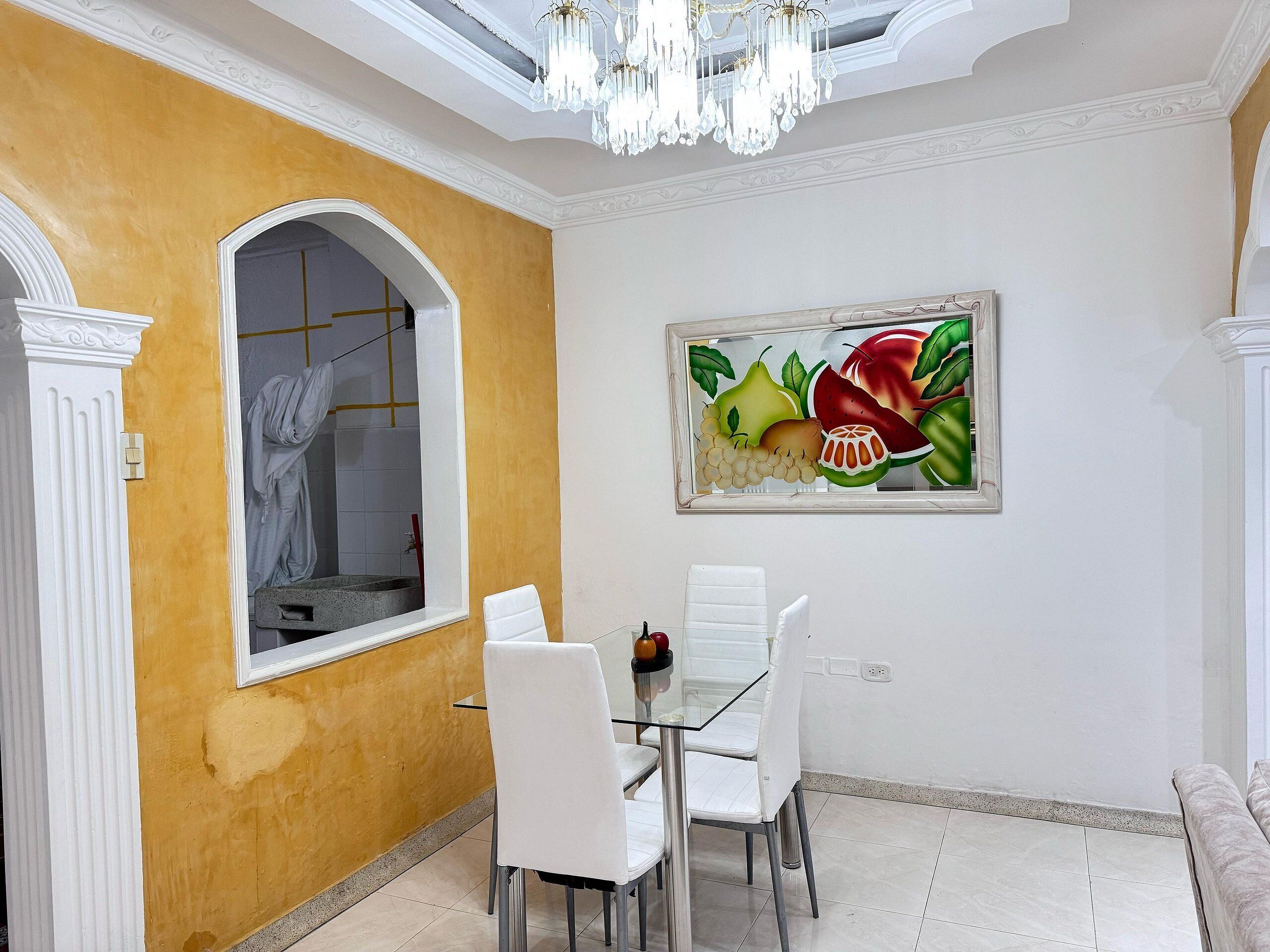 Uma sala de jantar com mesa e cadeiras, um lustre e um quadro de frutas na parede.