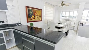 A modern kitchen with a black countertop, white cabinetry, a dining area with a white table and chairs, and a large window with a view. - Apartamento en Valledupar (Valledupar)