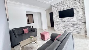 A modern living room with a grey sofa, a small coffee table, a pink ottoman, and a stone-textured wall with a mounted TV. - Apartamento en Valledupar (Valledupar)