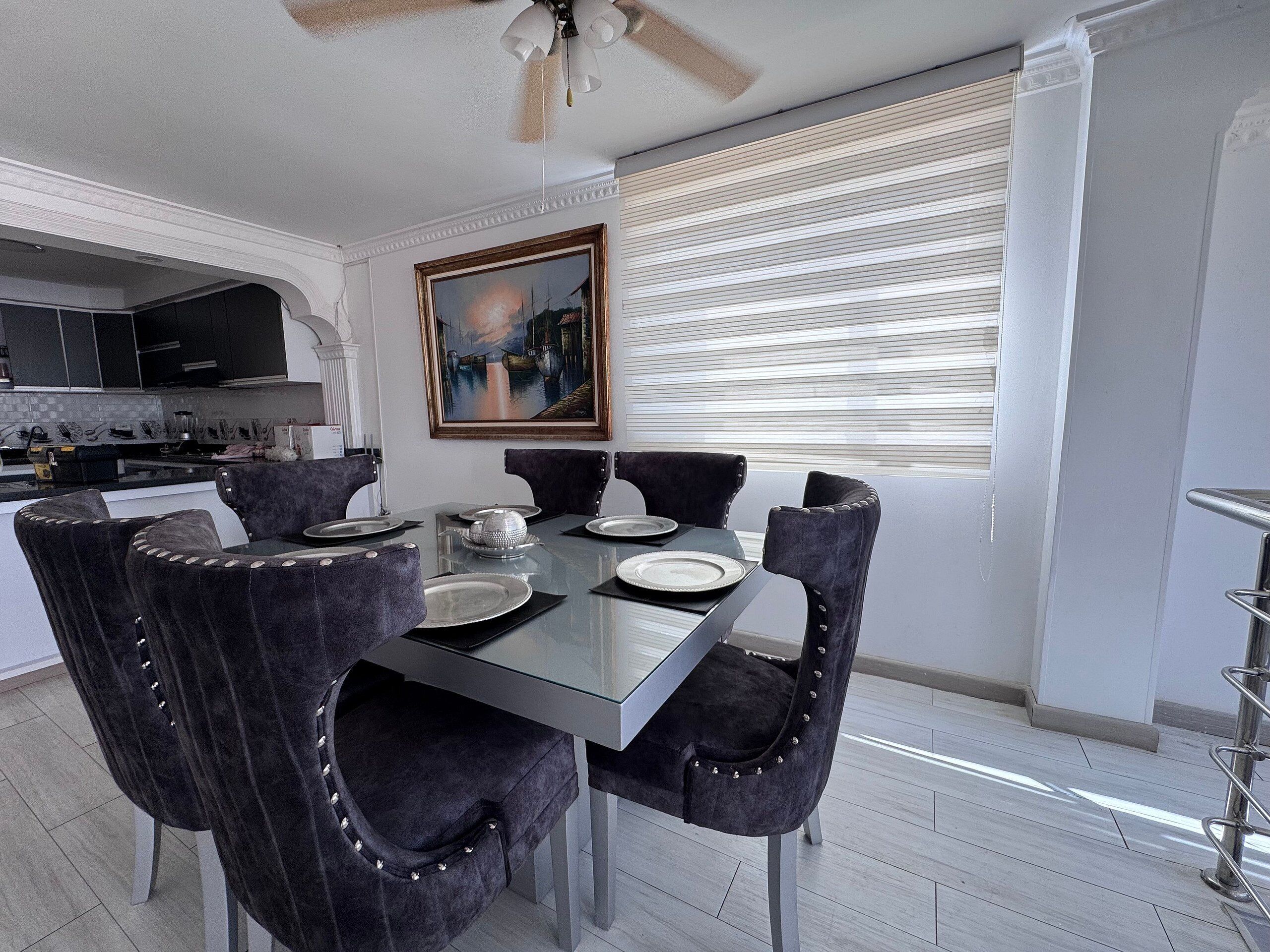 A dining area with a glass table, black upholstered chairs, a painting above the table, and a kitchen in the background.
