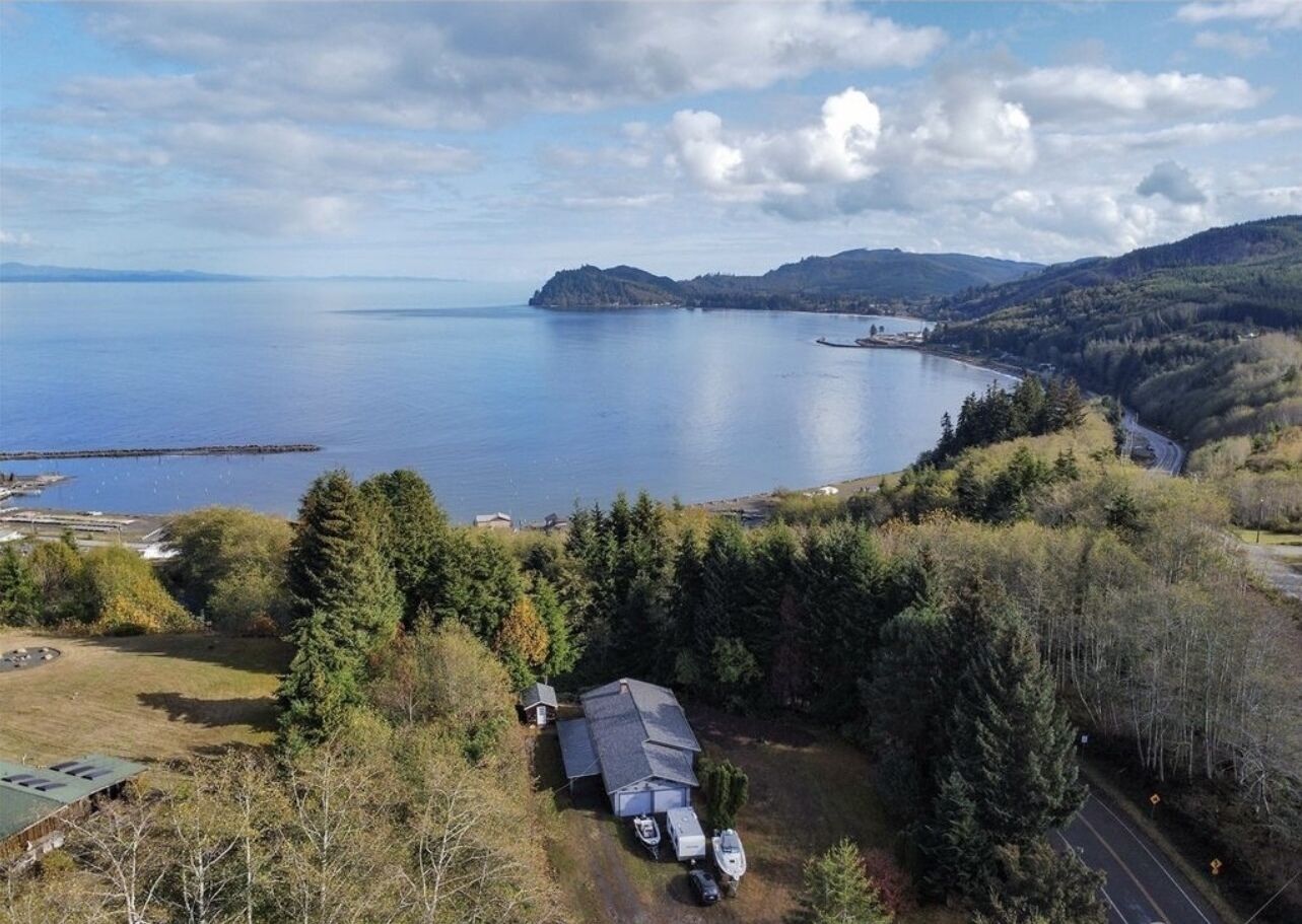 Une maison avec un grand garage, un bateau amarré à l’extérieur, et une vue sur un lac et des montagnes en arrière-plan.