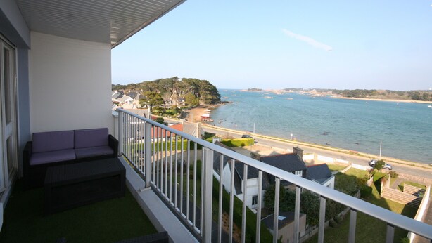 A balcony with a sofa and a small table, overlooking a coastal area with a beach and a forested hill. - Vue sur mer Epoustouflante (Trégastel)