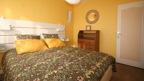 A bedroom with a floral bedspread, a white ceiling light, a wooden dresser, and a door. - Vue sur mer Epoustouflante (Trégastel)