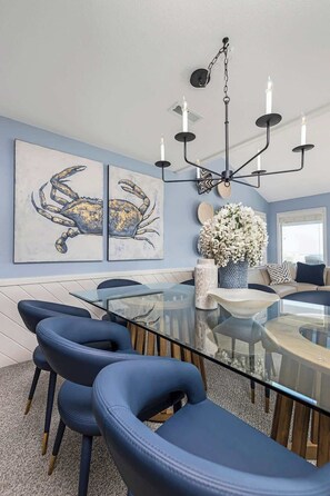 A modern dining area with a glass table, blue upholstered chairs, and a chandelier. - Beachfront, Heated POOL, Golf Cart & EV Charging (Ocean Isle Beach)