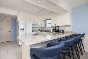 A modern kitchen with white cabinets, a granite countertop, a built-in microwave, a stove, and a coffee maker. There are four blue bar stools at the island. - Beachfront, Heated POOL, Golf Cart & EV Charging (Ocean Isle Beach)