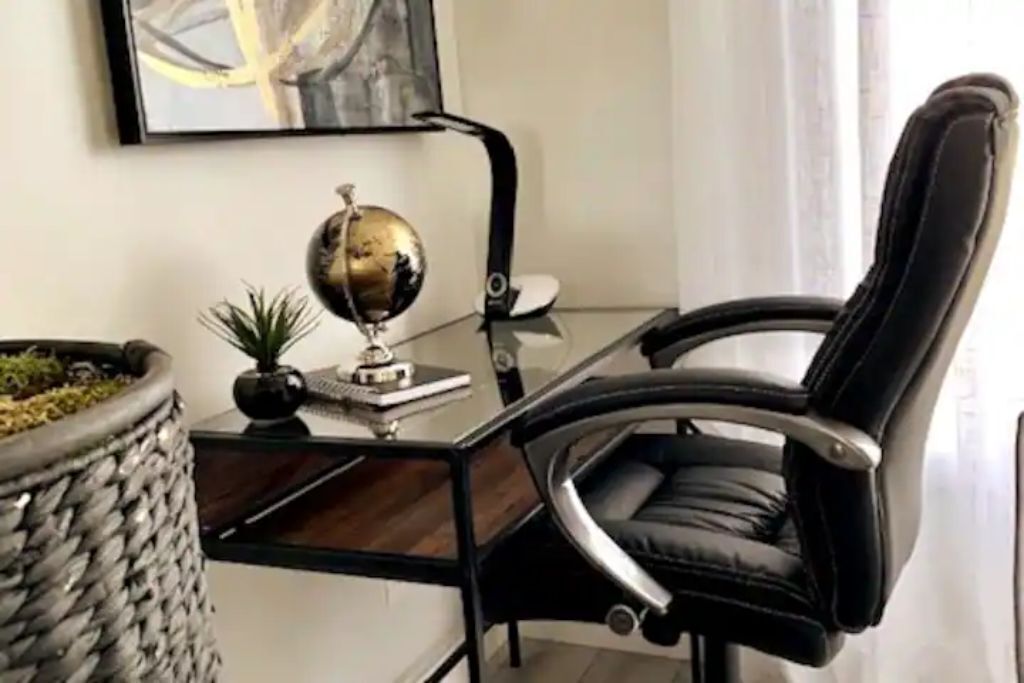 Een moderne bureaustoel met een hoge rugleuning en armleuningen, een glazen bureau met een zwart frame, een decoratieve wereldbol en een kleine potplant.