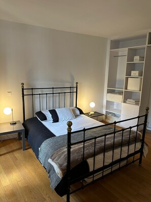 Ein ordentlich bezogenes Bett mit Metallgestell, zwei Nachttische mit Lampen und eine Einbauschränke mit Aufbewahrungsboxen.