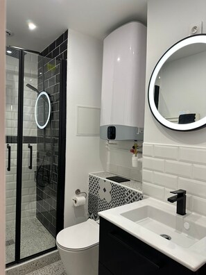 Ein modernes Badezimmer mit Duschkabine aus Glas, einer weißen Toilette, einem Waschbecken mit schwarzem Wasserhahn und einem Spiegel mit runder Beleuchtung.
