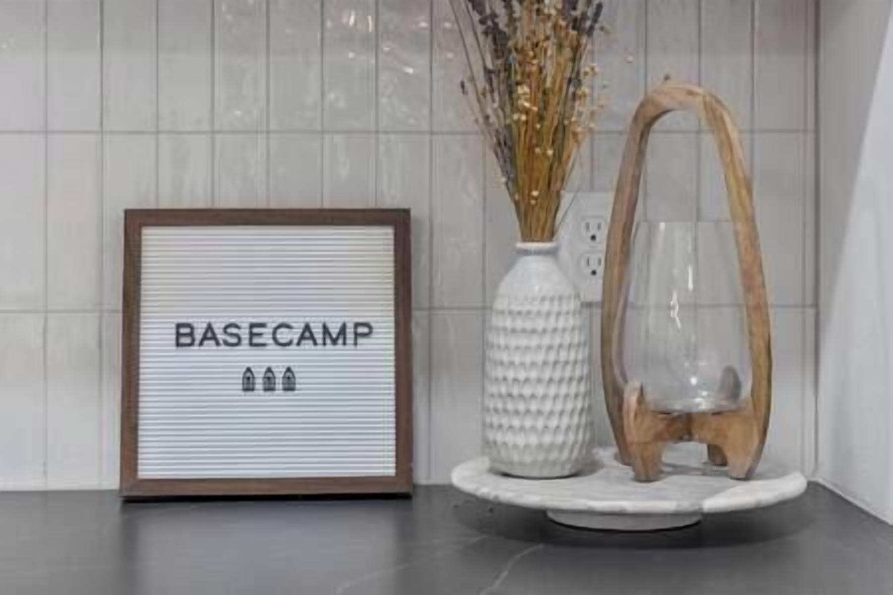 A framed sign with "BASECAMP" and three icons, a white vase with dried flowers, and a clear glass vase on a white pedestal.