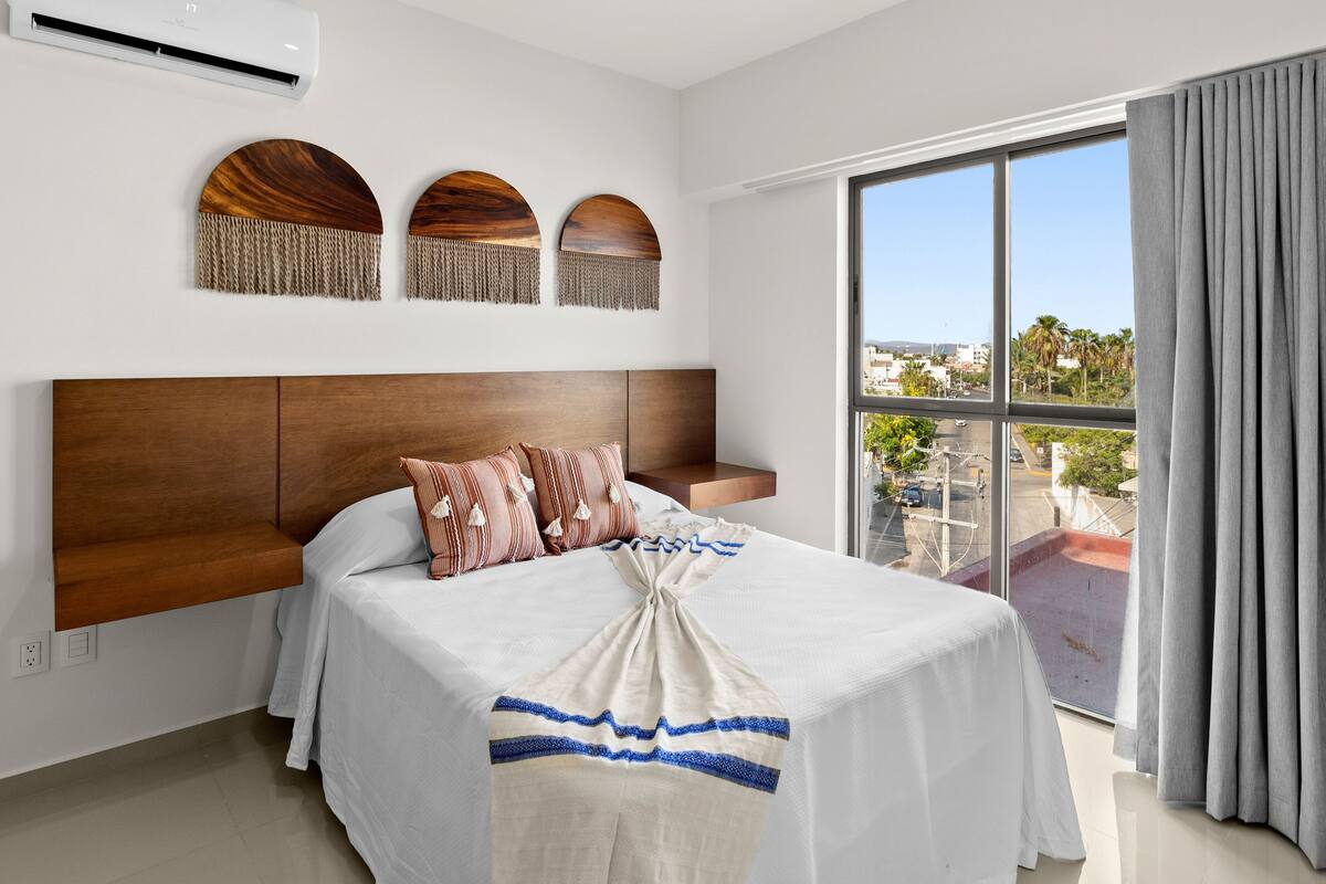 A hotel room with a large bed, a wooden headboard, a view of the city through a large window, and a balcony with a view of the street.