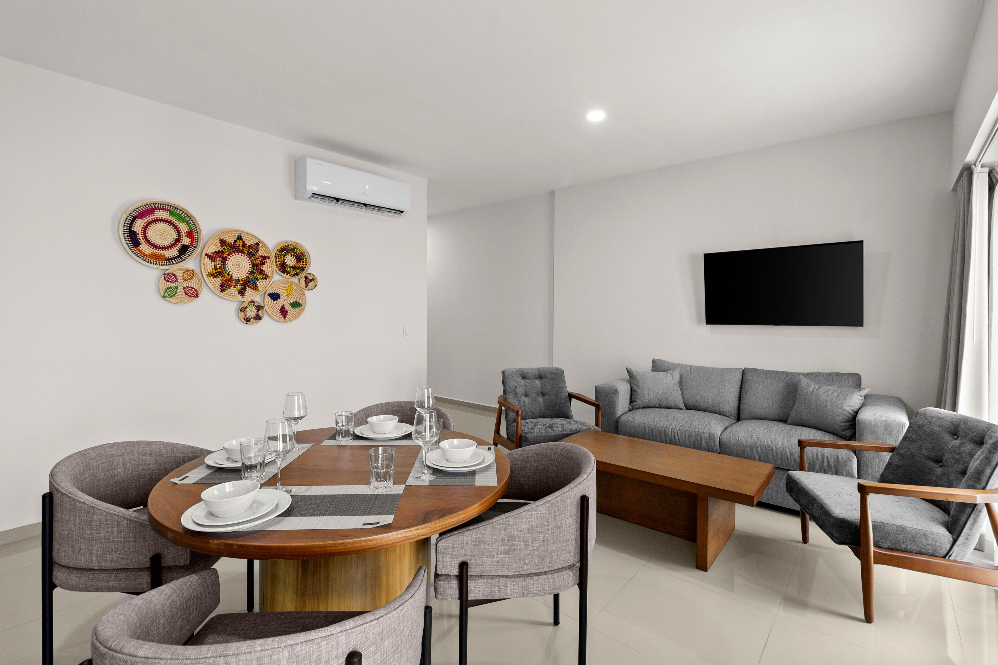 A modern living room with a round dining table set for four, grey upholstered chairs, a grey sofa, and a flat-screen TV mounted on the wall.