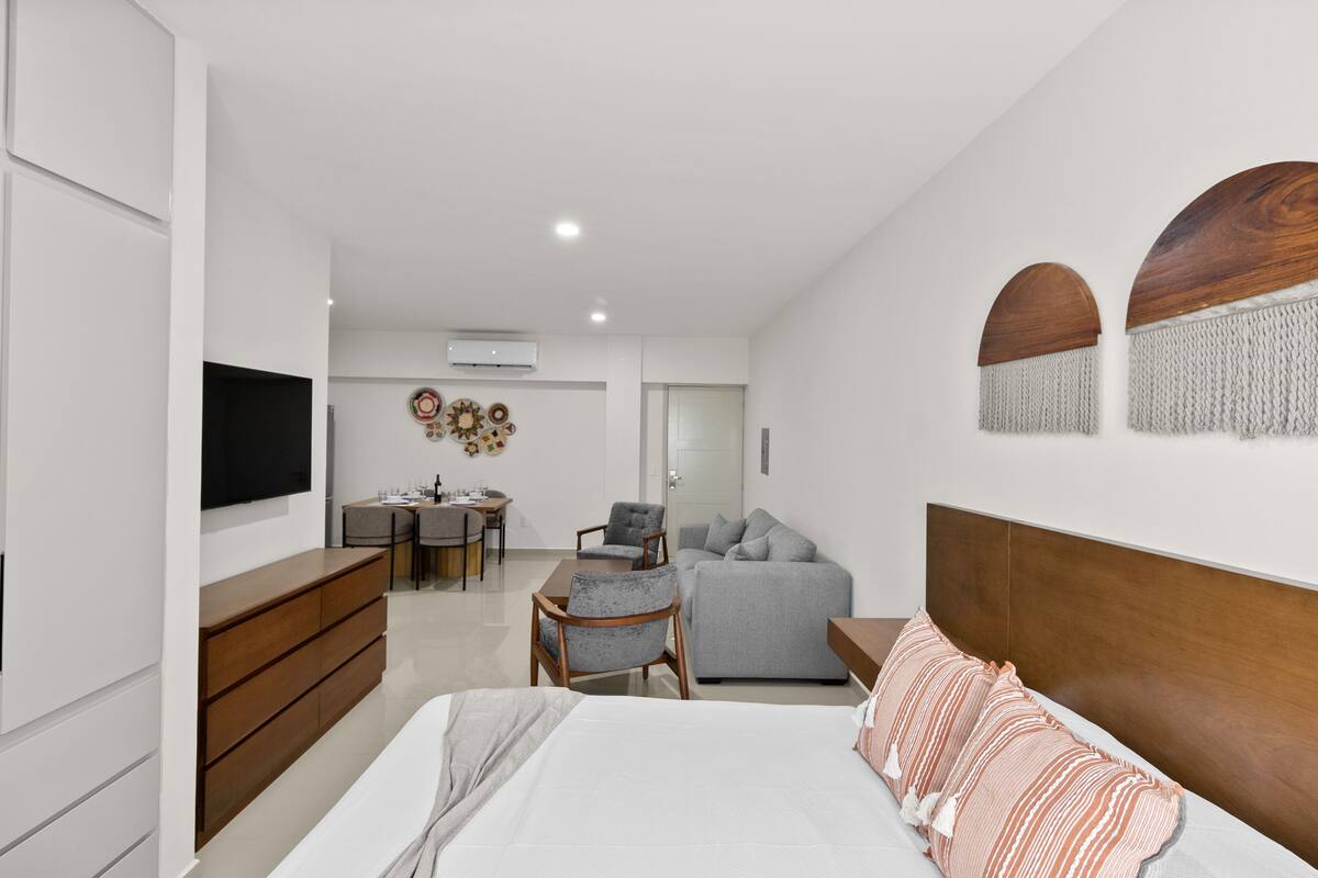 A modern bedroom with a bed, a grey sofa, a wooden dresser, a TV, and a desk with chairs.