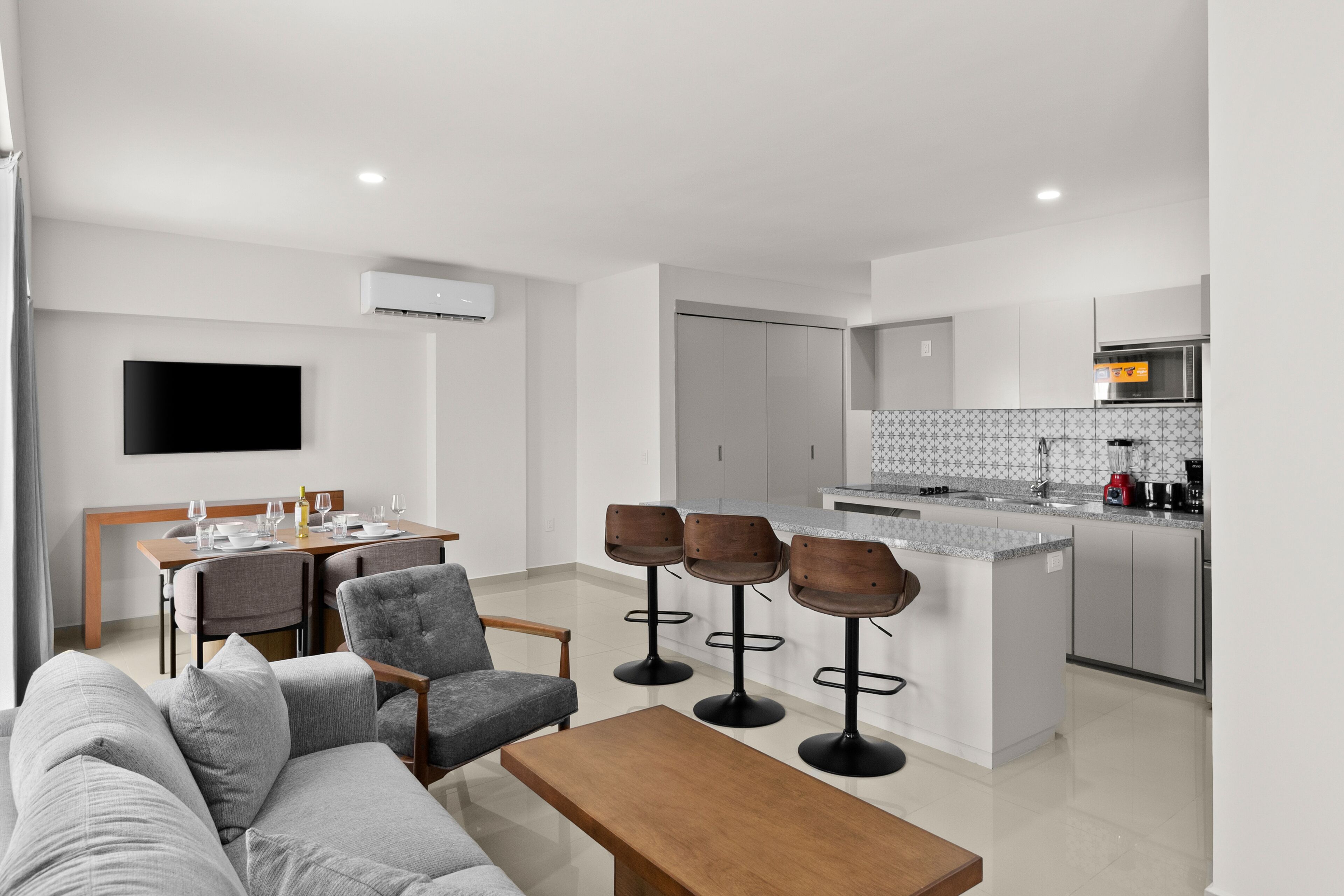 Un salon moderne avec un canapé, une table basse et des tabourets de bar. On aperçoit un coin repas avec une table et des chaises. Au fond, on distingue une cuisine équipée d’un four à micro-ondes et d’un évier.