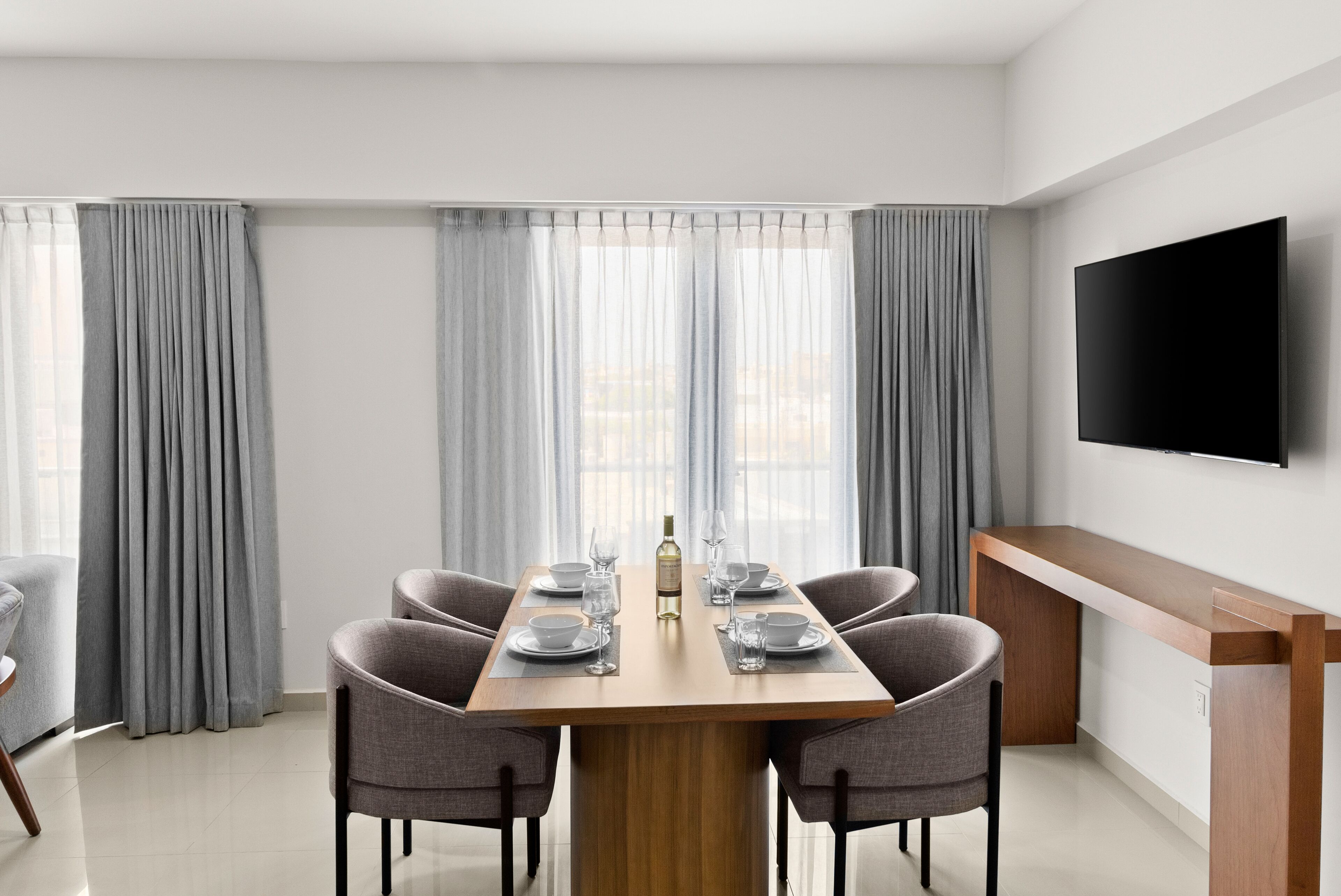Un comedor moderno con una mesa de madera dispuesta para cuatro, cortinas grises y un televisor de pantalla plana en la pared.
