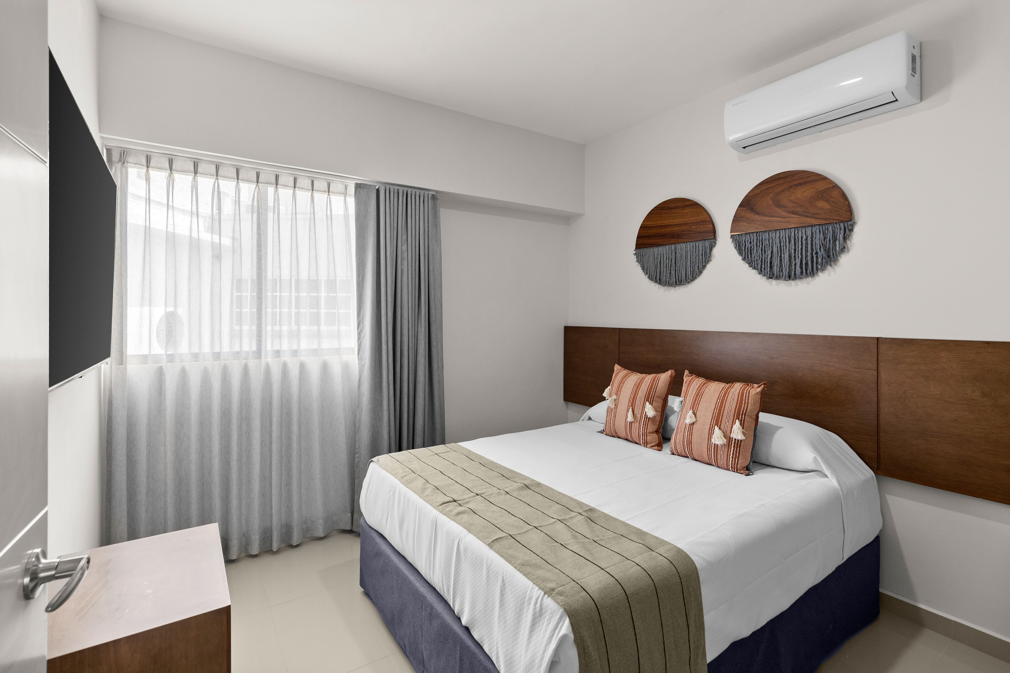 A modern hotel room with a large bed, a wooden headboard, a flat-screen TV, a built-in desk, and a window with curtains.
