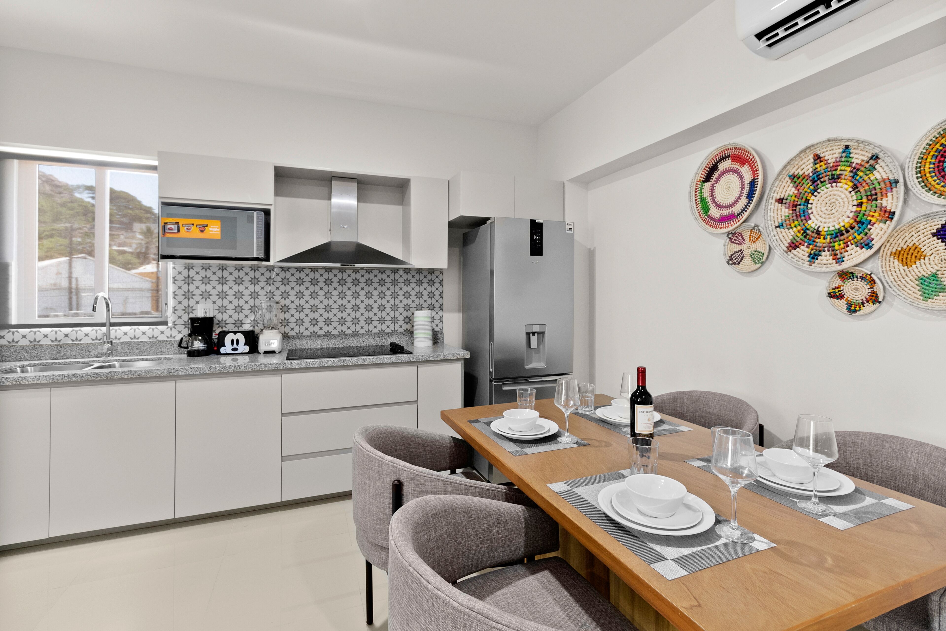 A modern kitchen with white cabinets, a microwave, and a refrigerator. There is a dining table set with plates and glasses.