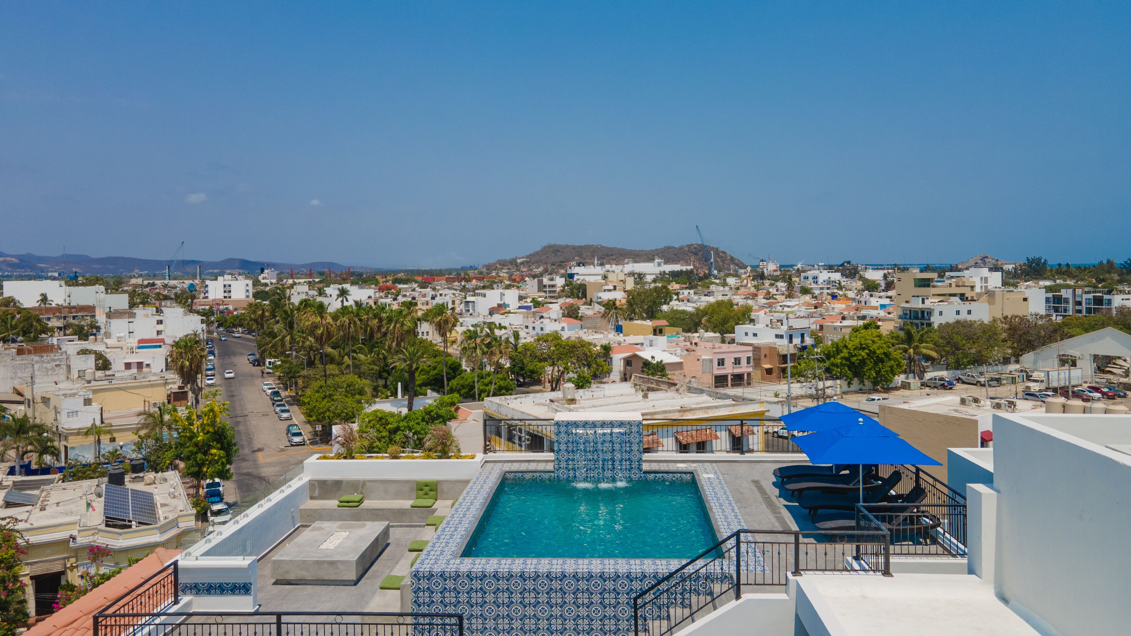 Una piscina en la azotea con diseño de azulejos azules y blancos, rodeada por un paisaje urbano con edificios y palmeras.