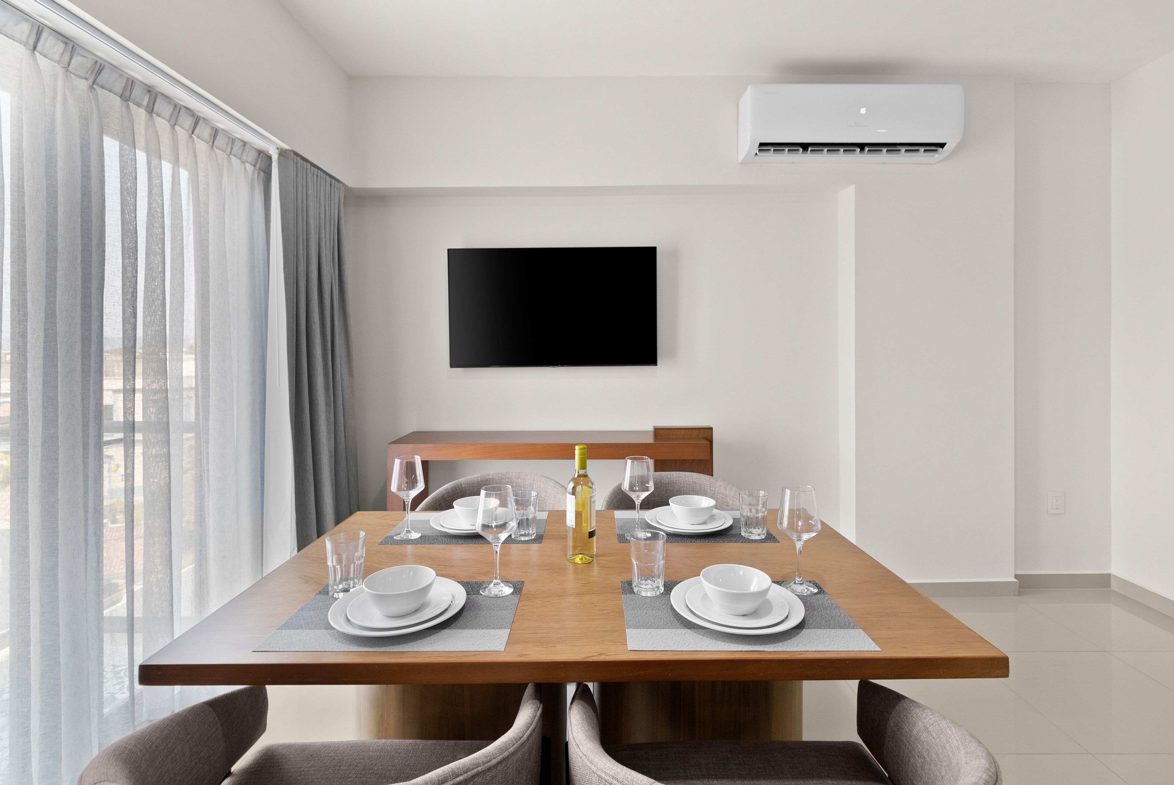 A modern dining area with a wooden table set for four, a flat-screen TV mounted on the wall, and an air conditioning unit above.