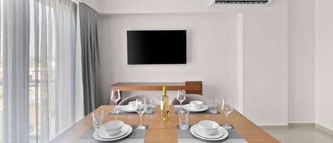 A modern dining area with a wooden table set for four, a flat-screen TV mounted on the wall, and an air conditioning unit above.