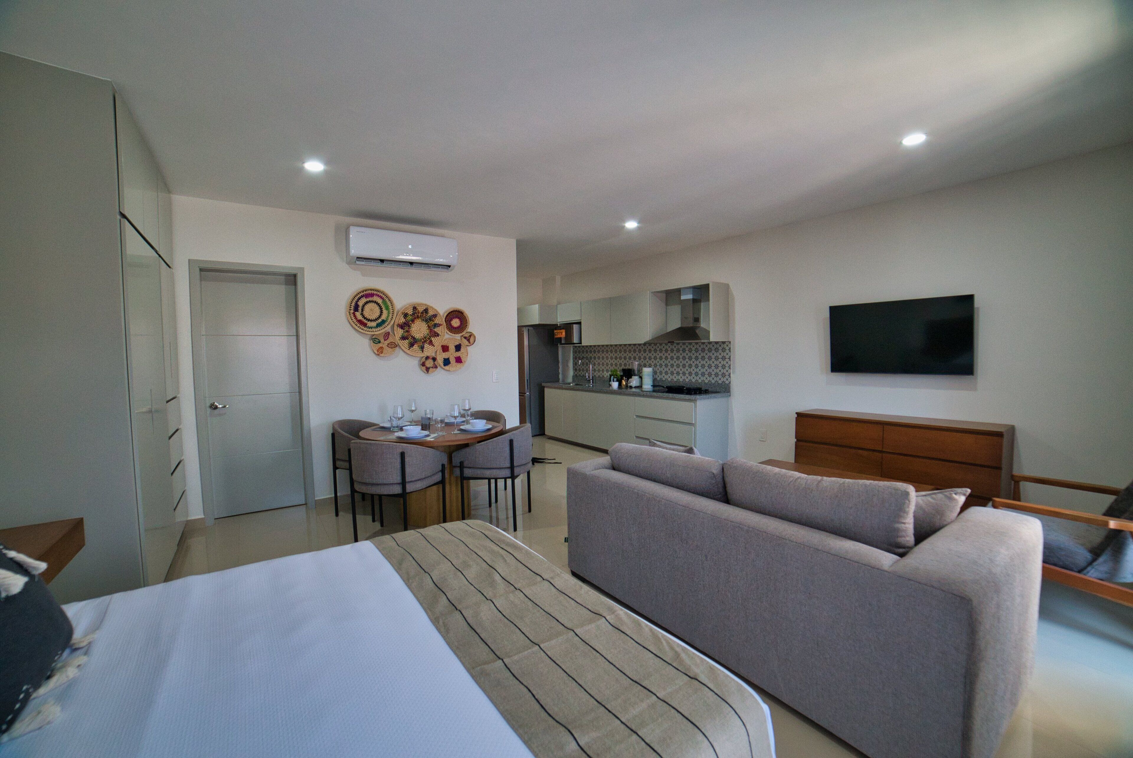A modern hotel room with a bed, a sofa, a dining area with a table and chairs, a kitchen area, and a TV mounted on the wall.