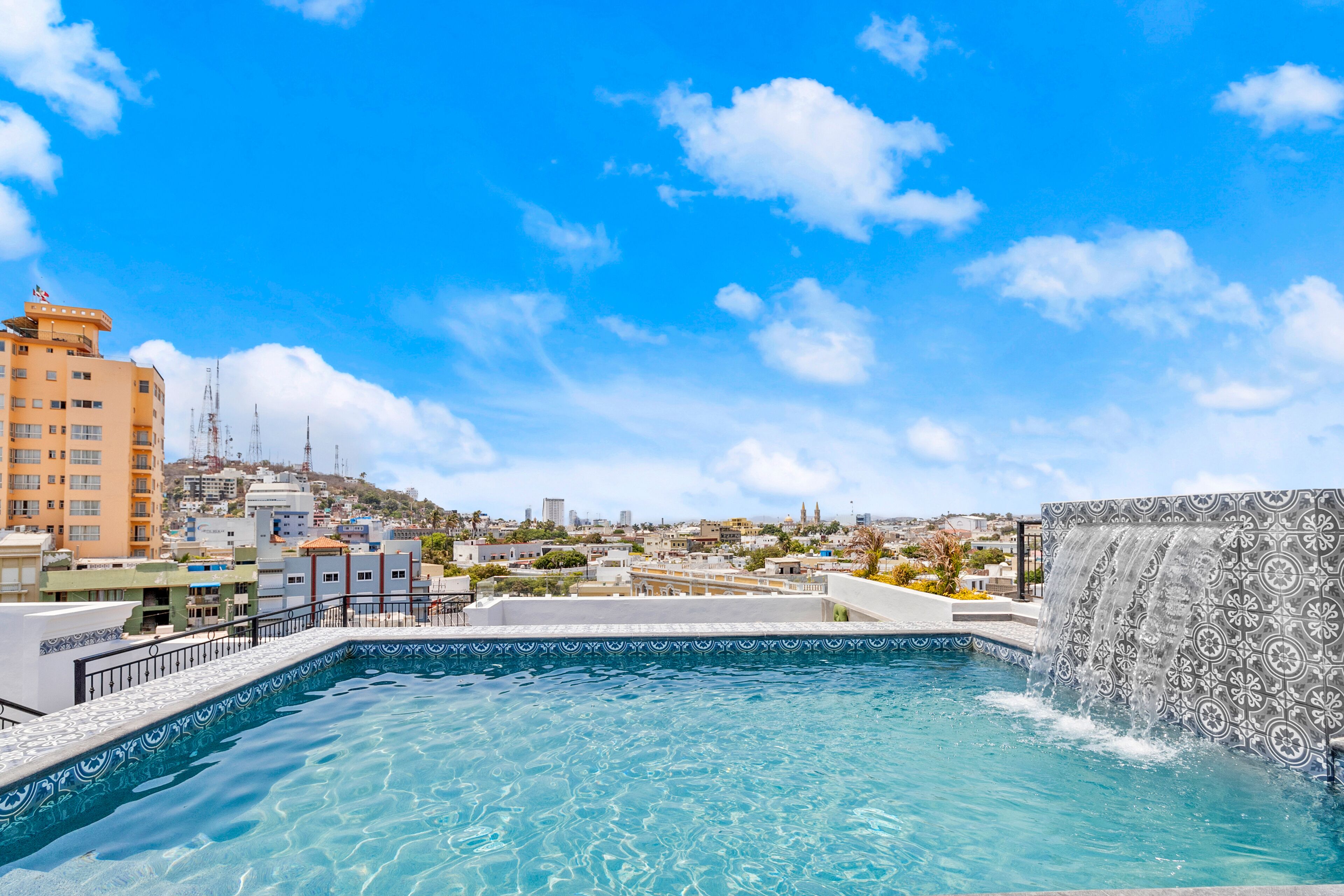 A rooftop pool with a waterfall feature, surrounded by city buildings and a clear blue sky.