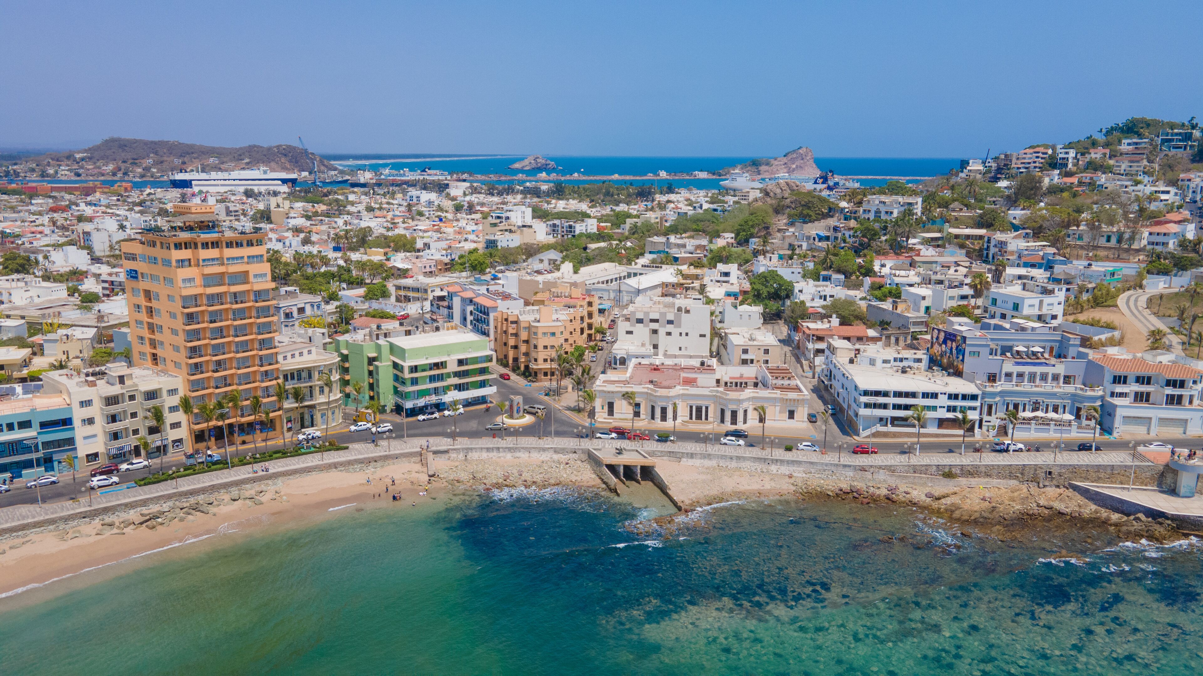 A coastal cityscape with a prominent high-rise hotel, a beach, and clear blue waters.