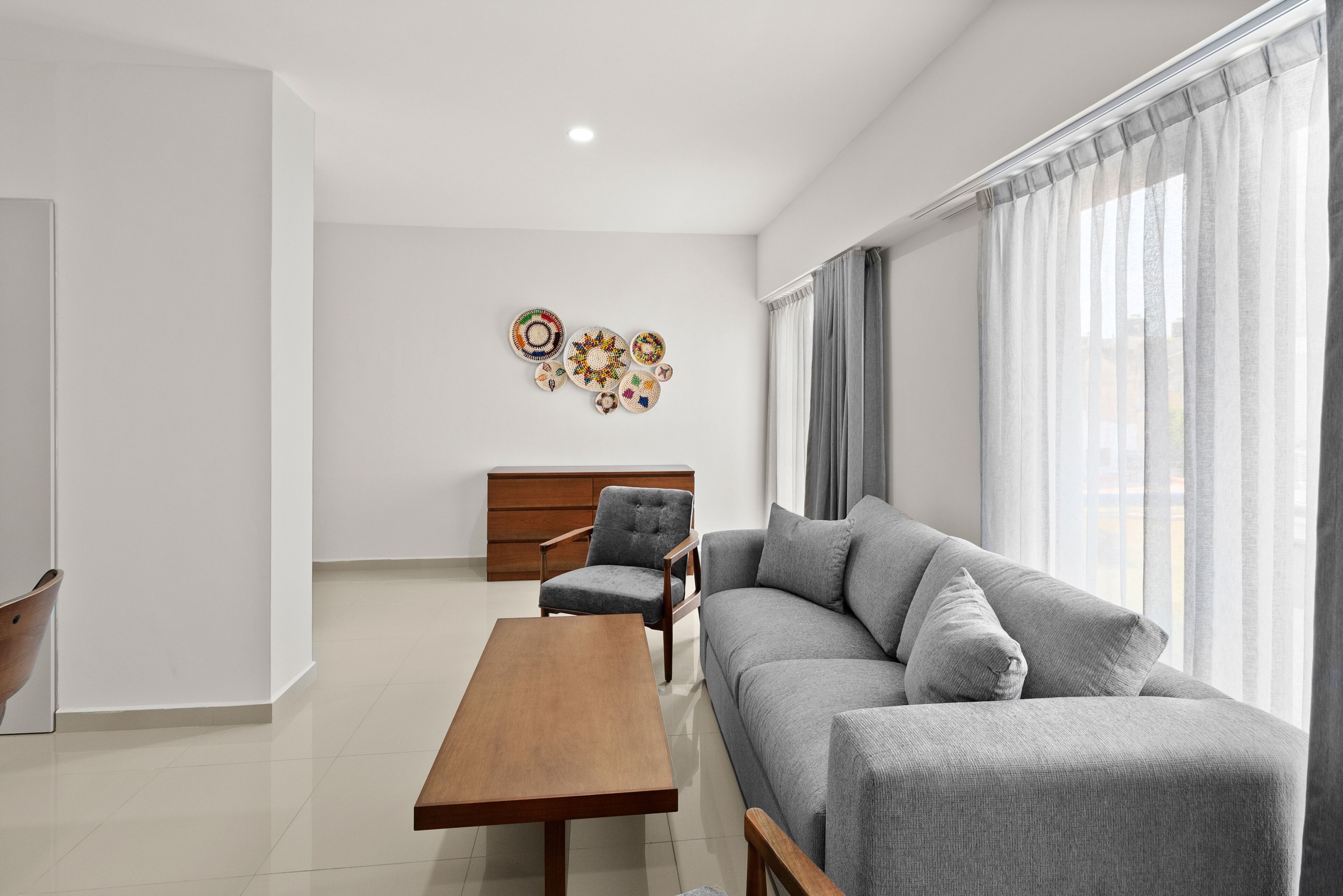 A modern living room with a grey sofa, a wooden coffee table, and a grey armchair. There is a wooden cabinet in the background and a wall with decorative plates.