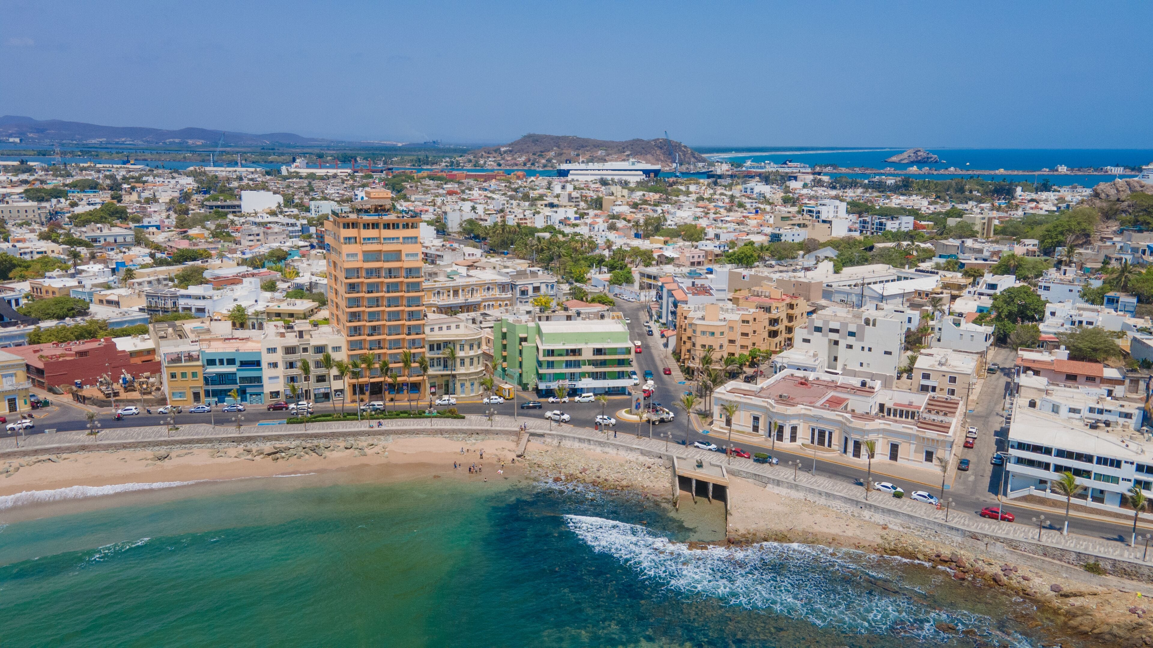 A coastal cityscape with a prominent hotel featuring a central courtyard, a beach, and a view of the ocean.