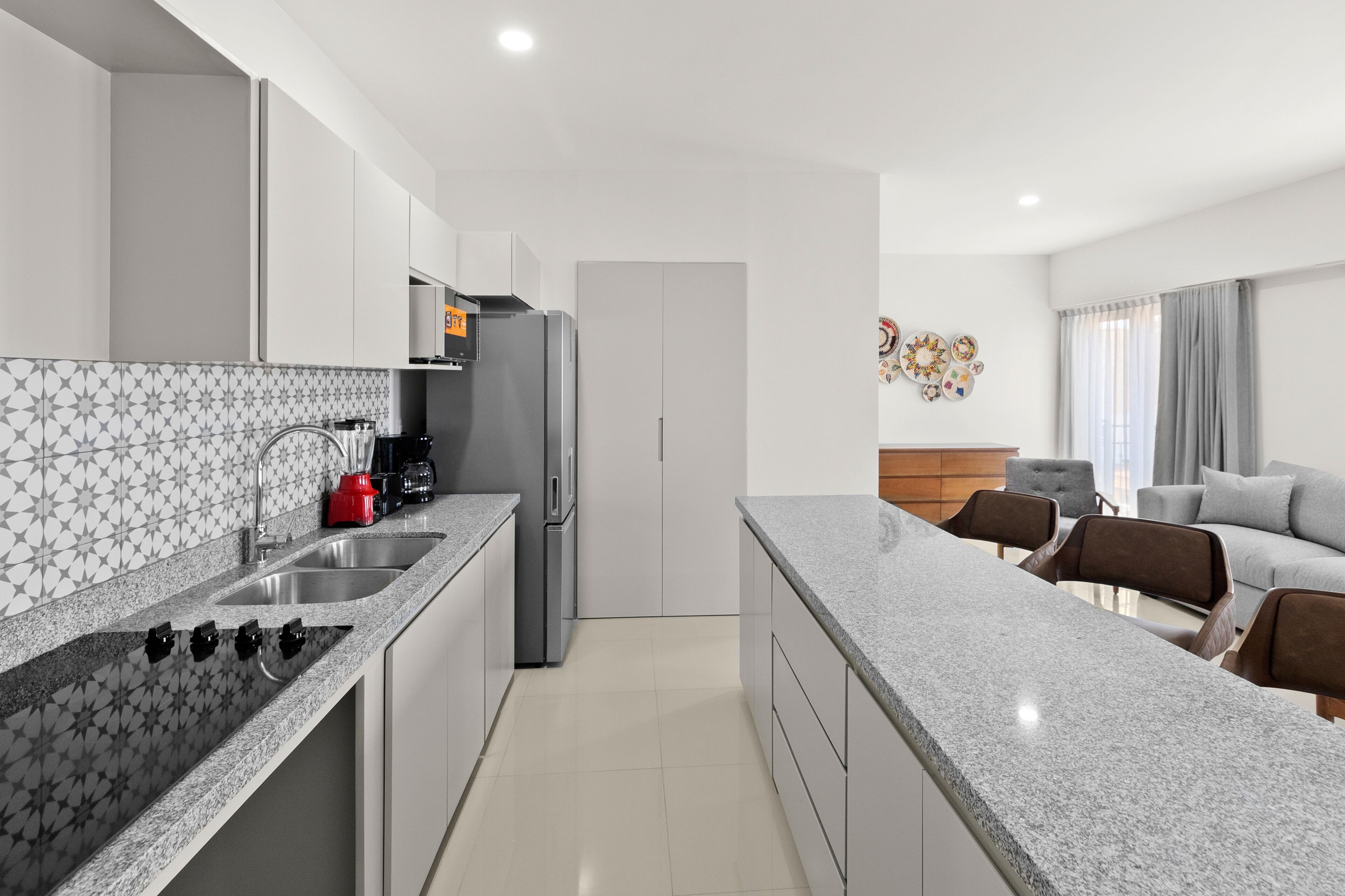 A modern kitchen with granite countertops, a built-in oven, and a refrigerator. There is a dining area with a gray sofa and a coffee table.