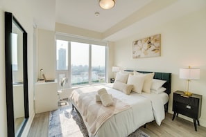 A modern bedroom with a large bed, two bedside tables, a window with a city view, and a painting on the wall. - Stylish Open Concept Condo w/Gym (Kitchener)