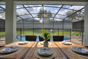 A covered patio with a table set for two, overlooking a swimming pool and a grassy area.