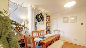 A dining area with a wooden table, chairs, a bench, a lamp, a wreath, and shelves with bottles and picture frames.