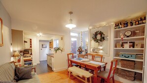 A cozy living room with a wooden dining table, a bench, and a sofa. There is a kitchen area in the background with a microwave and a refrigerator.