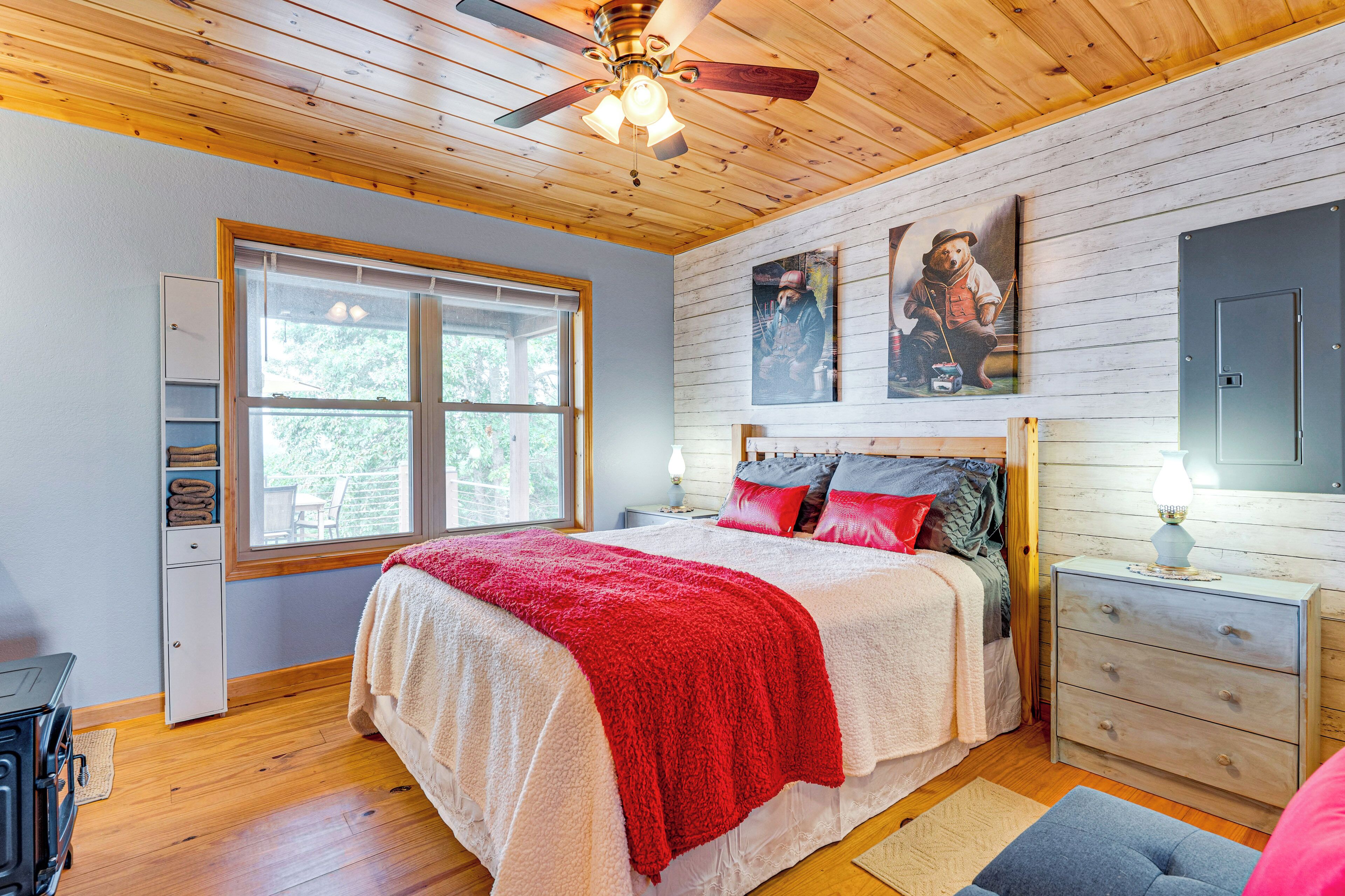 Une chambre avec un plafond en bois, un lit avec une literie rouge et blanche, une table de chevet avec une lampe et une fenêtre donnant sur des arbres.