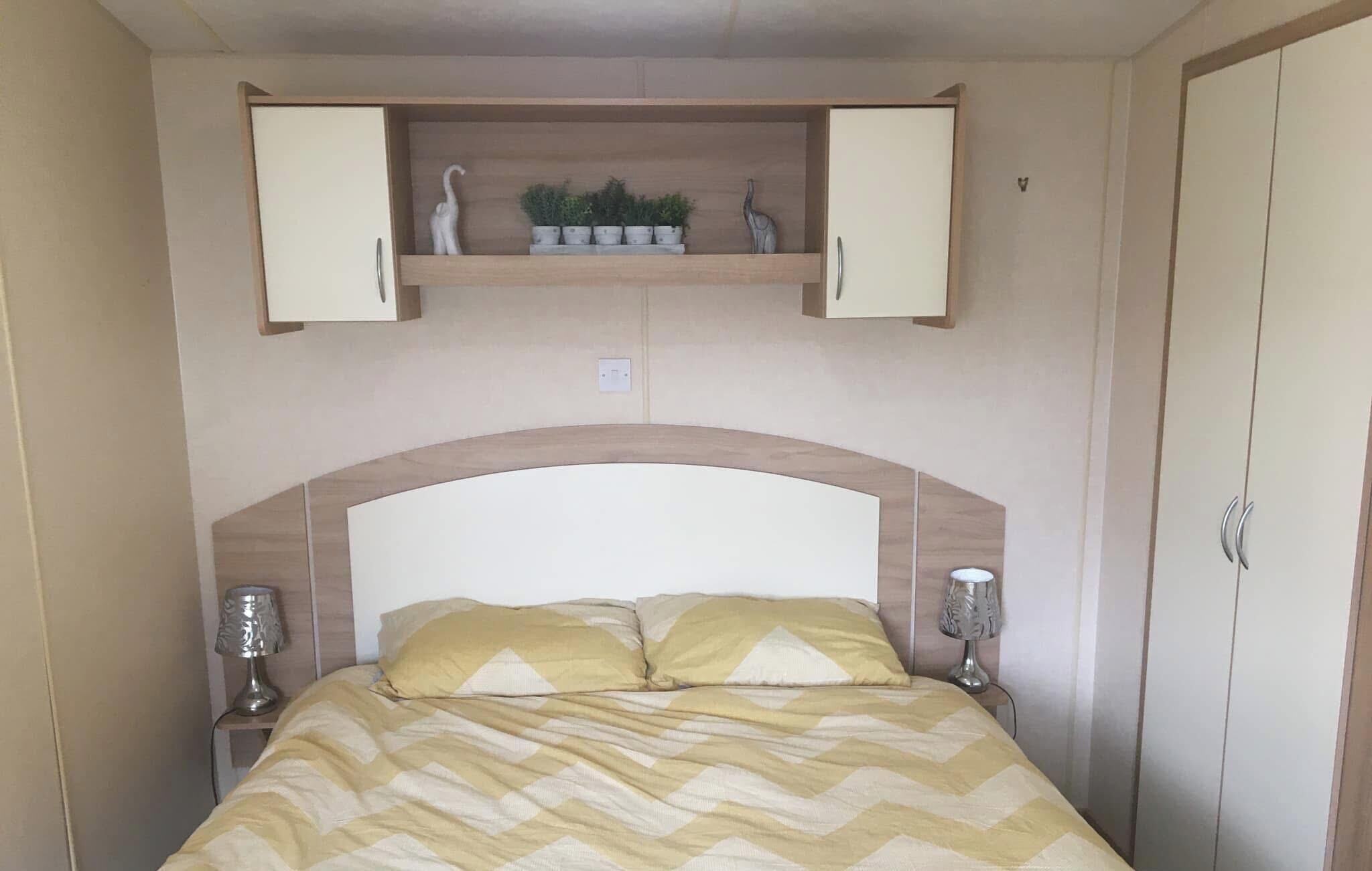 A bedroom with a bed, two bedside lamps, a wooden headboard, a shelf above the bed with decorative items, and a wardrobe.