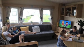 Une famille est réunie dans le salon, où se trouvent un canapé, un téléviseur et une table autour de laquelle les enfants sont en train de dessiner.