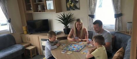 Una familia jugando un juego de mesa en una mesa de madera en una sala de estar con una televisión, estanterías y una planta en maceta.