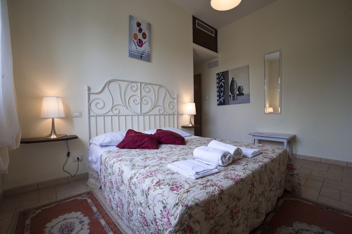 Un letto rifatto con cura, coperto da lenzuola a motivi floreali, due cuscini e asciugamani arrotolati. Due lampade da comodino, un tavolino e uno specchio fissato alla parete.
