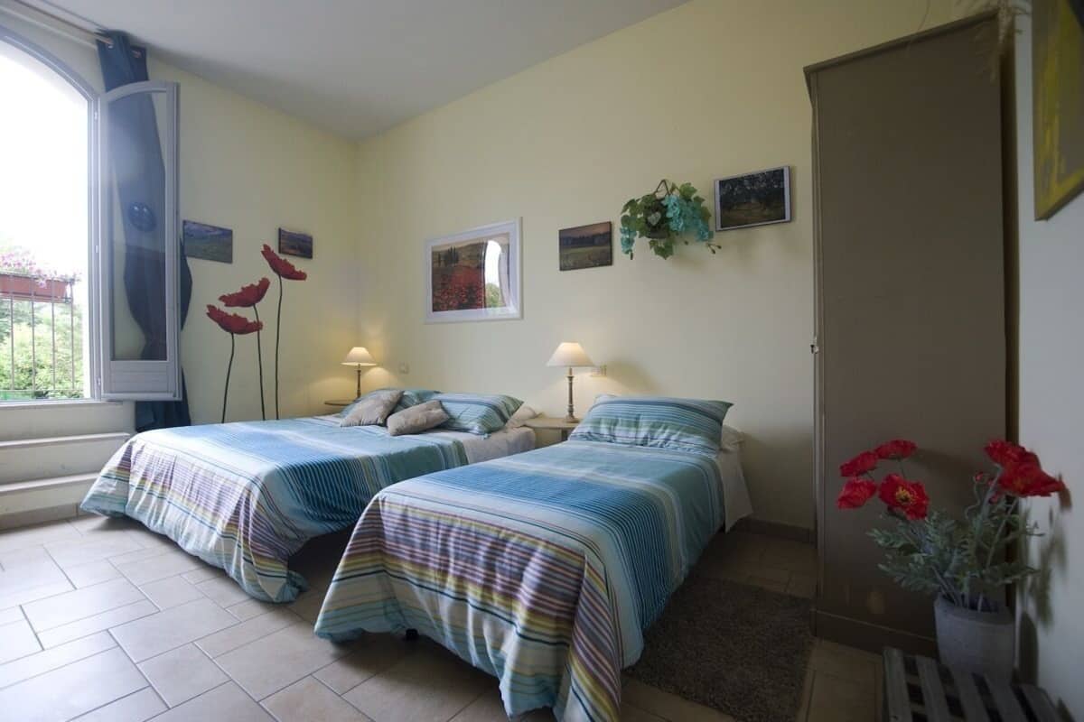 Une chambre avec deux lits, une fenêtre avec des rideaux, une porte et un mur orné de tableaux et d’une plante en pot.