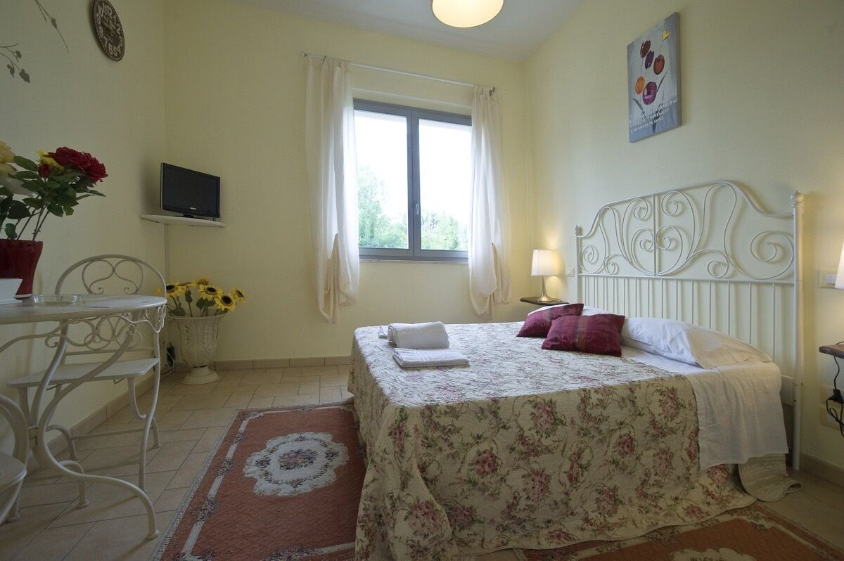 Una camera da letto con un letto, una TV, una finestra con tende, un tavolino con un vaso di fiori e una sveglia a parete.