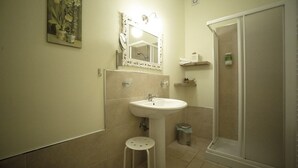 Un baño con lavamanos pedestal, un espejo grande con iluminación, cabina de ducha y un taburete pequeño.
