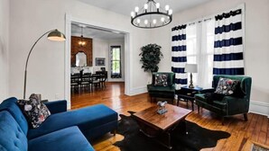 Ein Wohnzimmer mit einem blauen Sofa, einem Holztisch und einer Kristallleuchter. Im Hintergrund ist ein Essbereich mit einer Ziegelwand und einer Pflanze zu sehen.