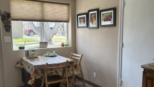 A dining area with a table covered in a floral tablecloth, wooden chairs, and a window with blinds. There are framed pictures on the wall and a door to the right.
