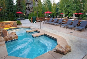 A hot tub area with lounge chairs and red umbrellas, surrounded by stone walls and trees. - Charlie Rentz: Arrowhead Ski-In/Out, Steps to Lift, Sleeps 8, Heated Pool + Hot Tub (Edwards)