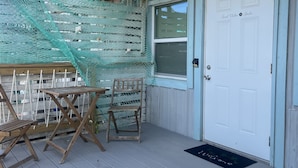A wooden deck with a table and chairs, a white door with a nameplate, and a blue mat at the entrance.